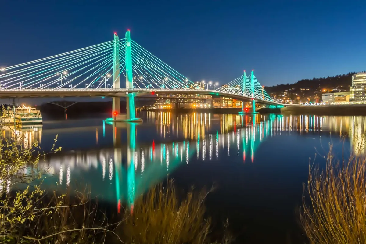 A scenic view of a suspension bridge reflecting in the water, illuminated by city lights at dusk, highlighting the tranquil yet lively character of the surrounding area.
