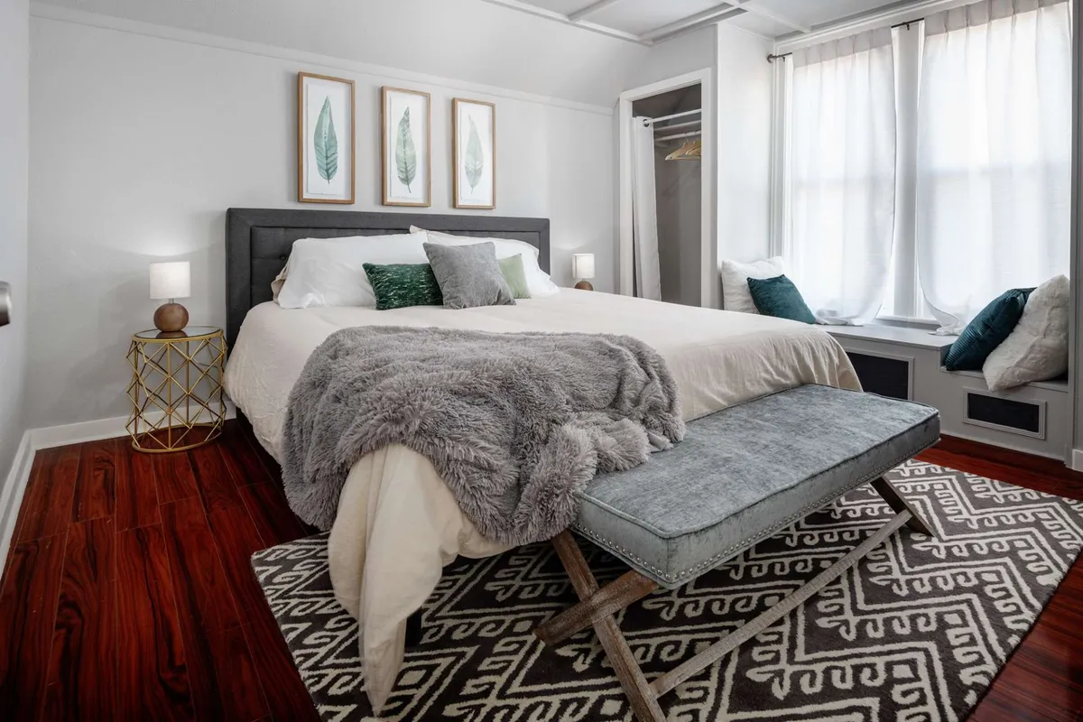 This image features a sunlit bedroom with a comfortable bed adorned with decorative pillows, a cozy throw blanket, and a stylish bench at the foot. Framed leaf artwork and light streaming through the curtains create a serene, inviting atmosphere ideal for relaxation.