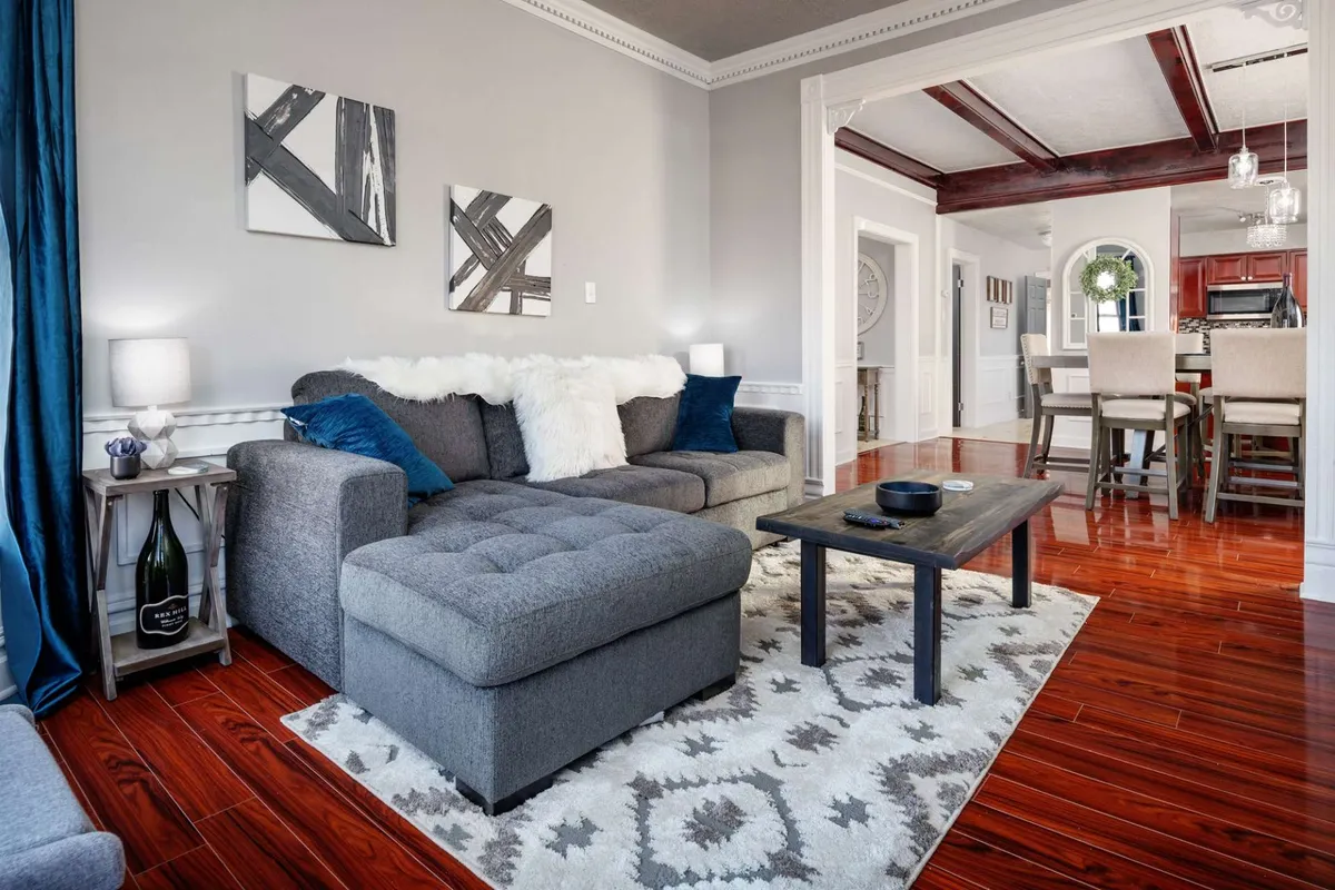 The image showcases a bright, inviting living room with a plush sofa, stylish coffee table, patterned area rug, and tasteful artwork. The seating arrangement encourages conversation and relaxation, while the decor blends Victorian charm with modern comfort. The space is warmly lit, creating an atmosphere ideal for gathering with friends or family.