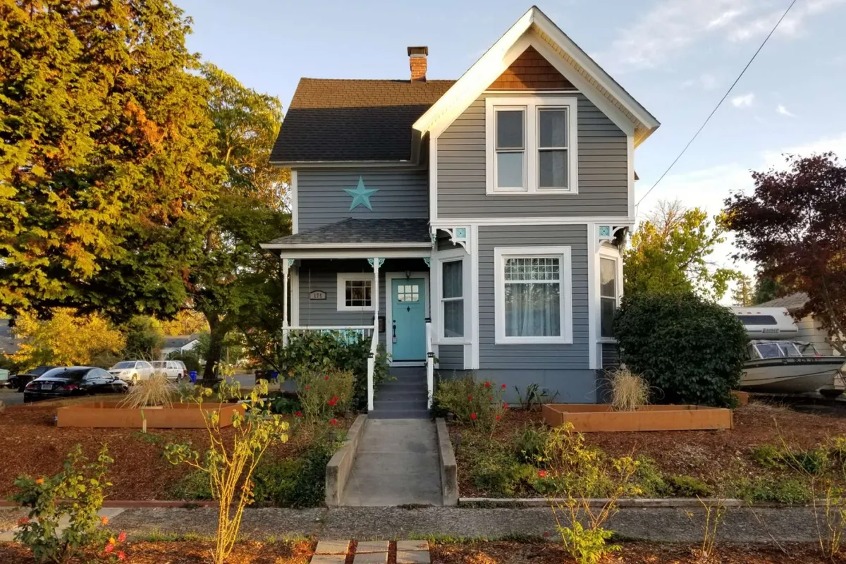 The exterior of a charming Victorian home featuring a blue front door, white trim, star accent on the facade, and blooming flower beds. The inviting porch is bathed in sunset light, suggesting a warm and welcoming setting for families.