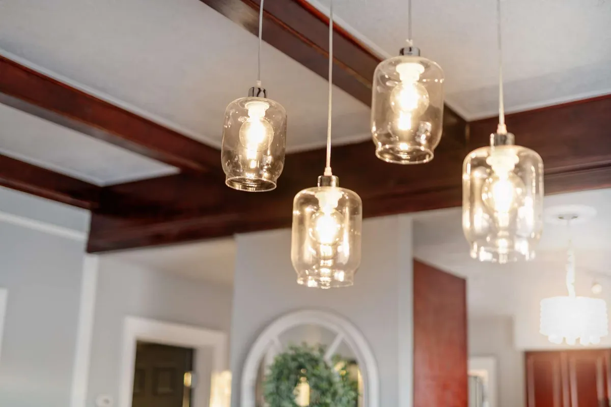 A cozy dining area featuring hanging glass light fixtures, wooden ceiling beams, and a decorative wreath on the wall. The setup radiates warmth, inviting guests to gather for meals and conversation.