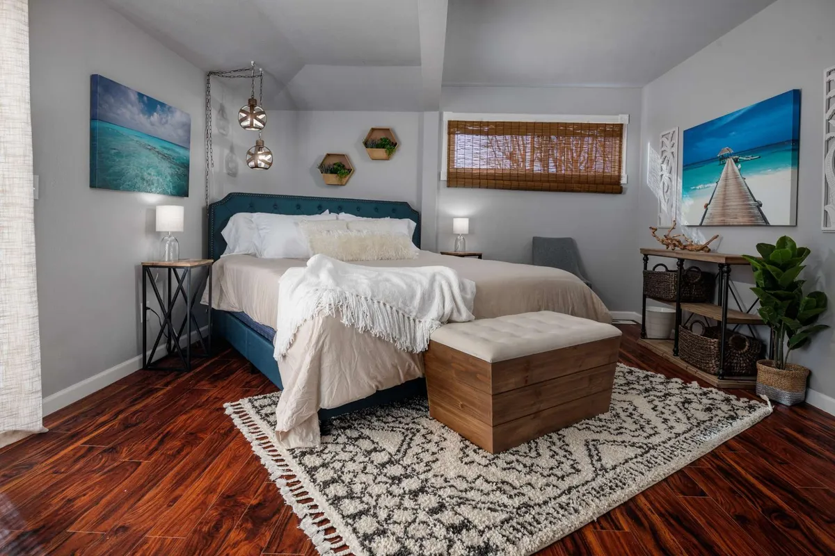 A calm, cozy bedroom highlighted by a blue headboard, decorative wall art, and a wooden storage bench. Stylish bedside lamps provide soft lighting, while natural light filters in, creating an inviting spot for relaxation and quiet moments.