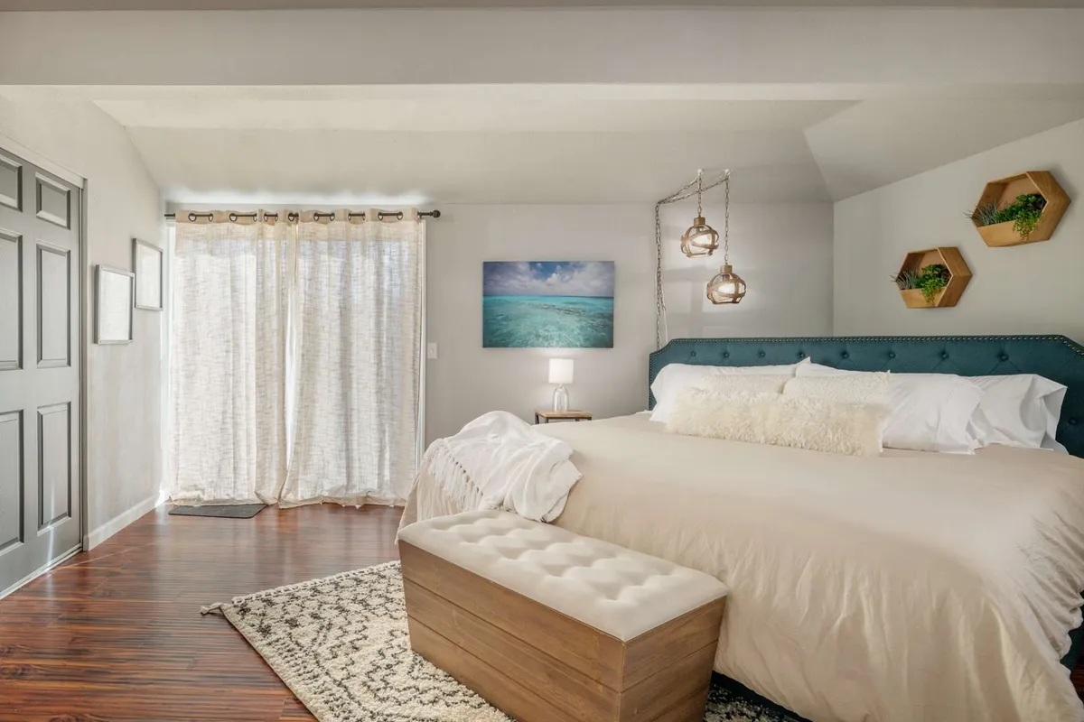 A spacious primary bedroom with a king-sized bed dressed in plush bedding, complemented by a decorative nightstand lamp, serene ocean artwork, hanging pendant lights, and a stylish wooden bench at the foot of the bed. Sunlight streams through the windows, illuminating the tranquil space and highlighting the inviting textures and calming atmosphere.