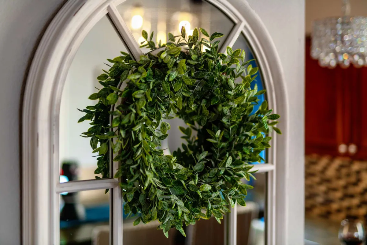 This image features a decorative wreath on the door, an arched mirror reflecting background furnishings, and subtle entryway decor, creating an inviting sense of arrival. The overall mood is warm and welcoming, suggesting a thoughtfully maintained historic home.