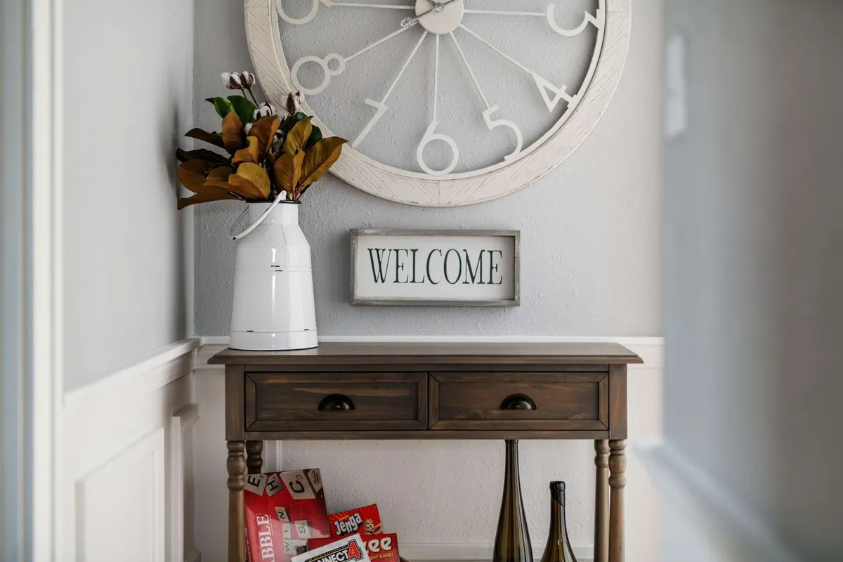 The scene showcases a large wall clock above a classic wooden console table, a welcome sign, decorative vase, and a stack of magazines. The space is styled to feel warm and homey, with elements that evoke history and hospitality.